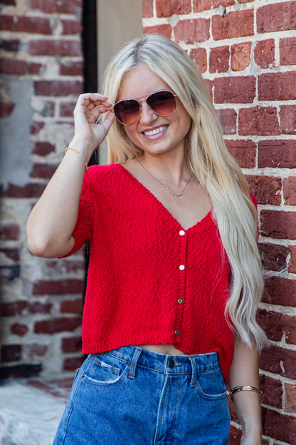 A smiling blonde woman wearing a bright red textured short-sleeve button-front cardigan and blue jeans, standing in front of a red brick wall.