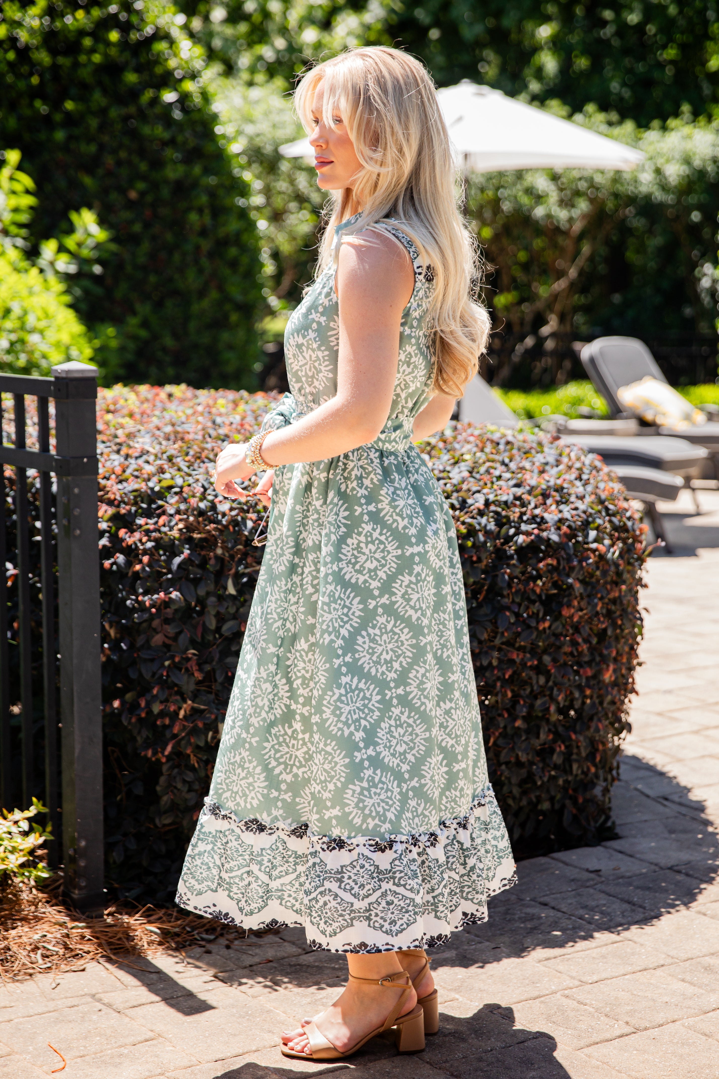 Woman in a floral dress standing outdoors near a bush and patio furniture.