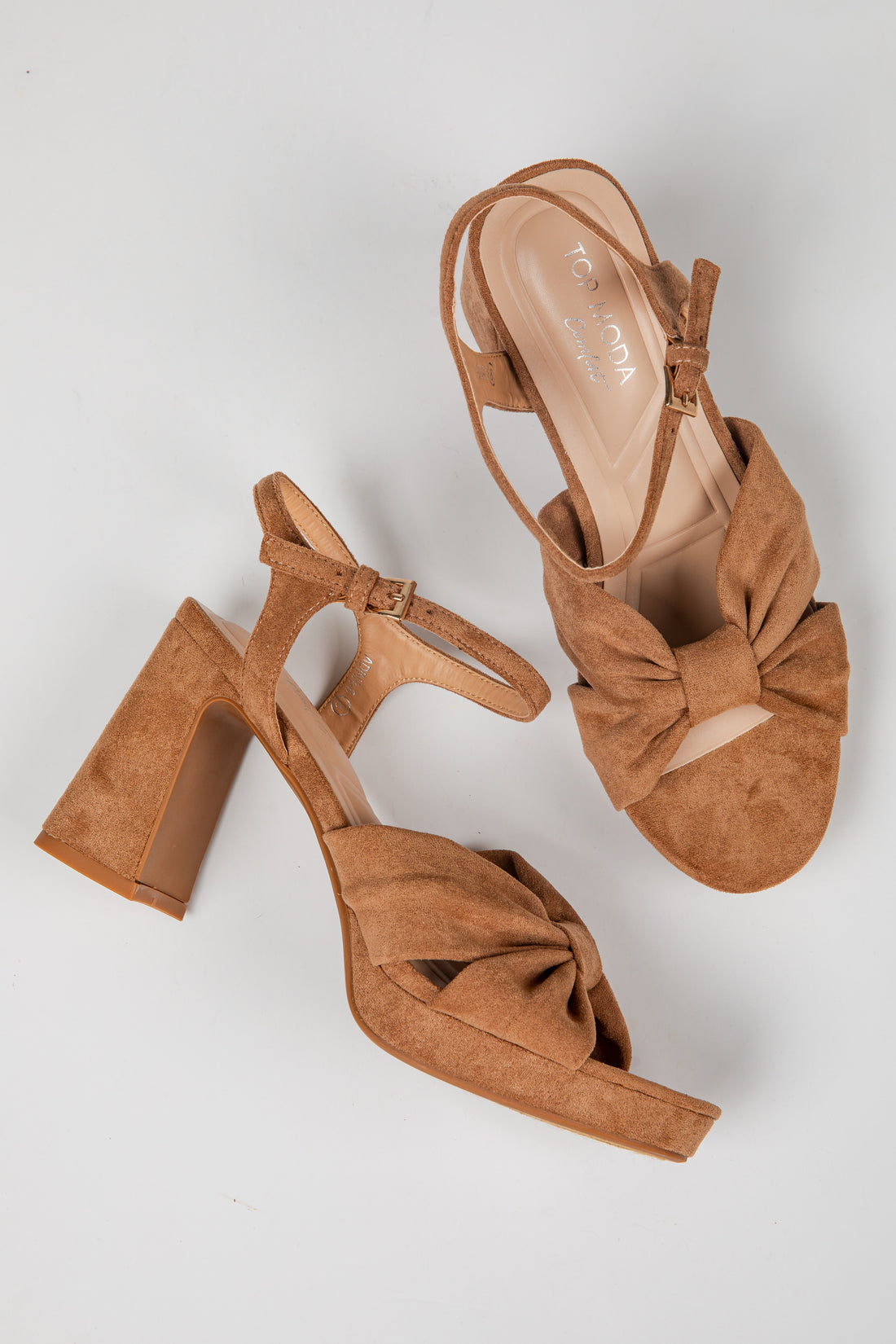 A pair of tan suede block-heel sandals with a large bow detail on the toe and ankle straps, displayed on a light gray background.