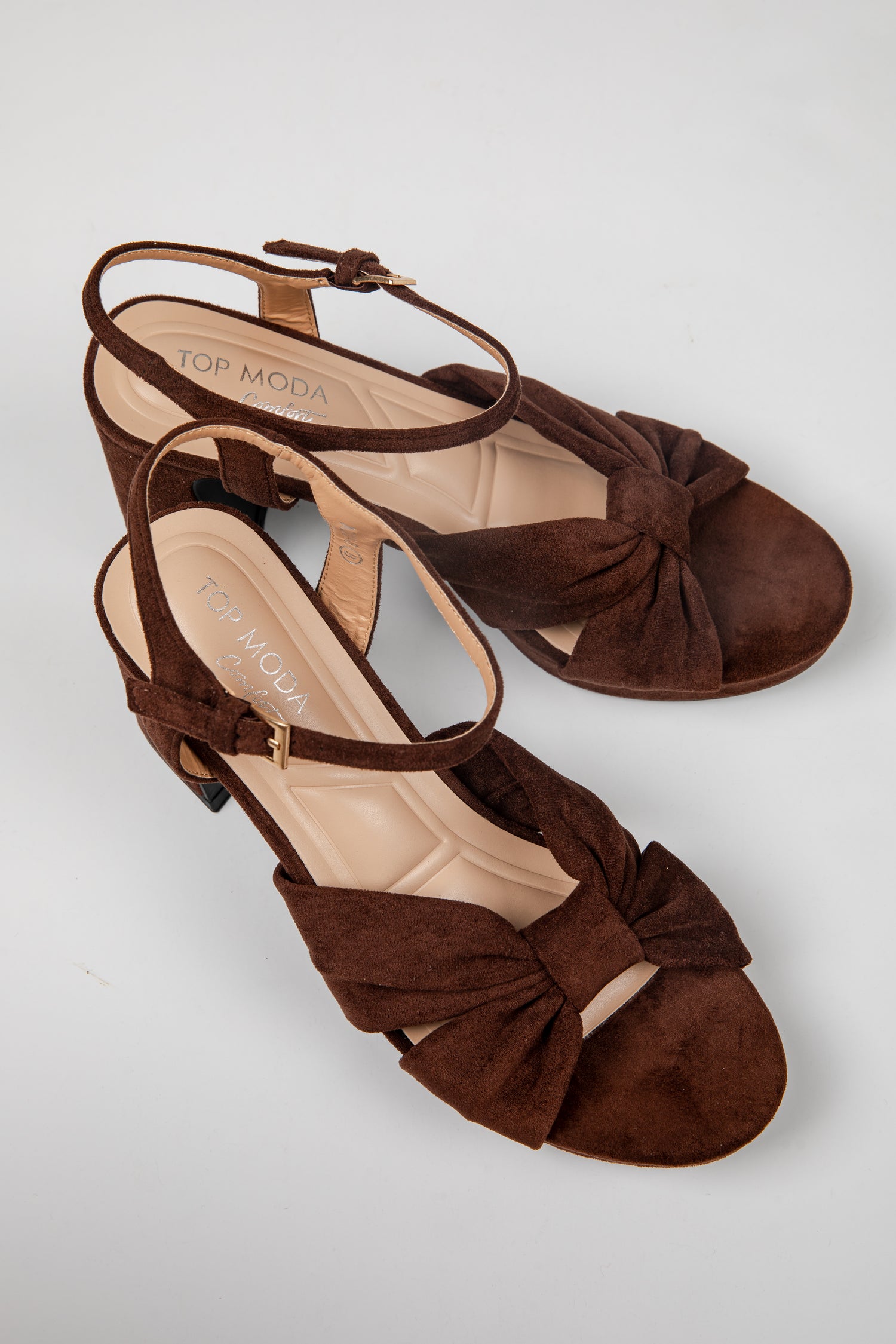 A pair of dark brown suede high-heeled sandals with bow details on the toes and ankle straps, displayed on a light gray background. The brand name &