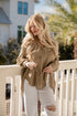 Woman standing on a balcony wearing a beige sweater and white pants, holding sunglasses.