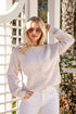 Woman wearing a light gray ribbed sweater and white pants standing in front of a decorative white lattice.