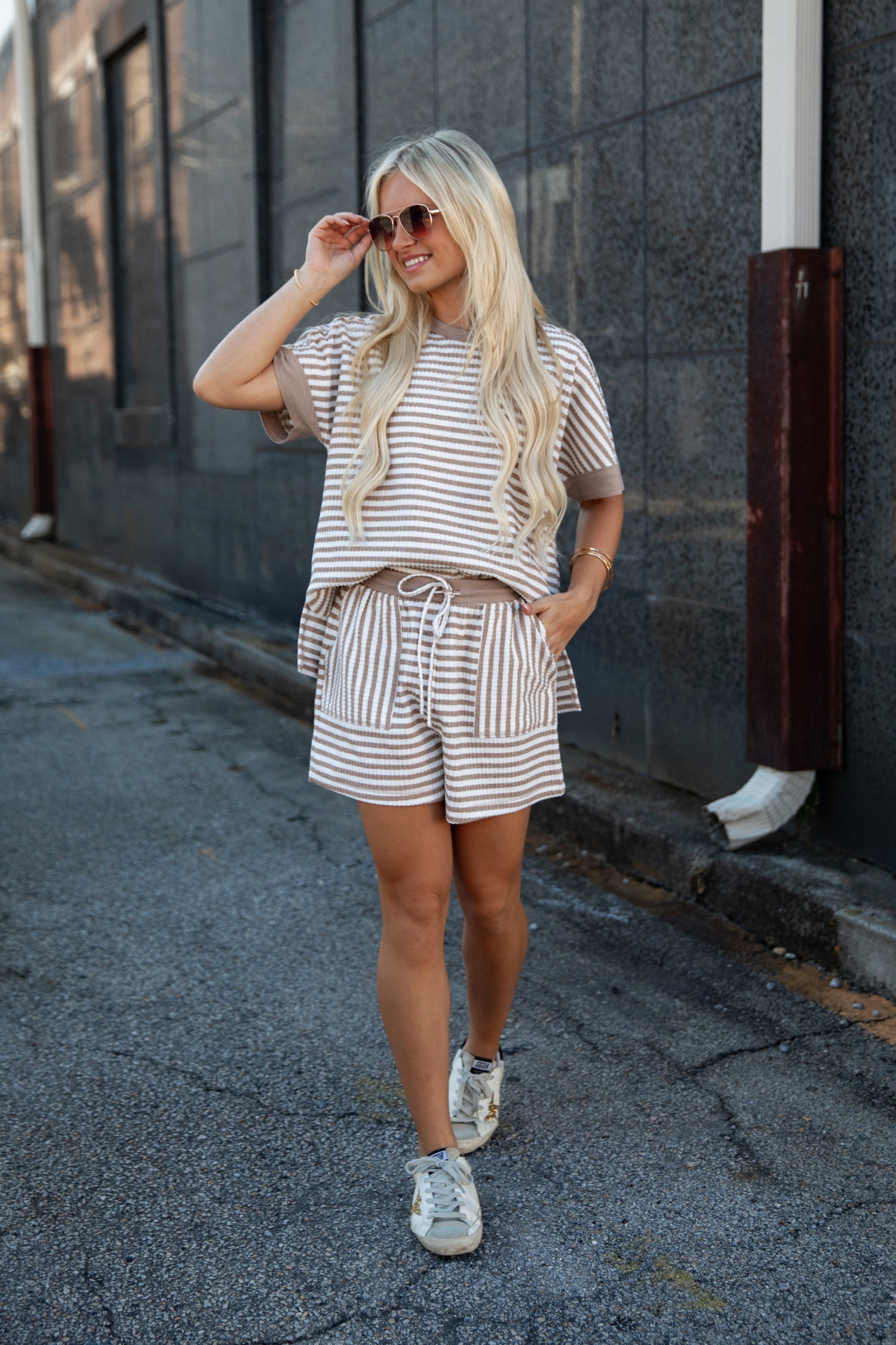 A woman wearing a matching beige and white striped lounge set with a short-sleeve top and shorts, walking down an outdoor alleyway.