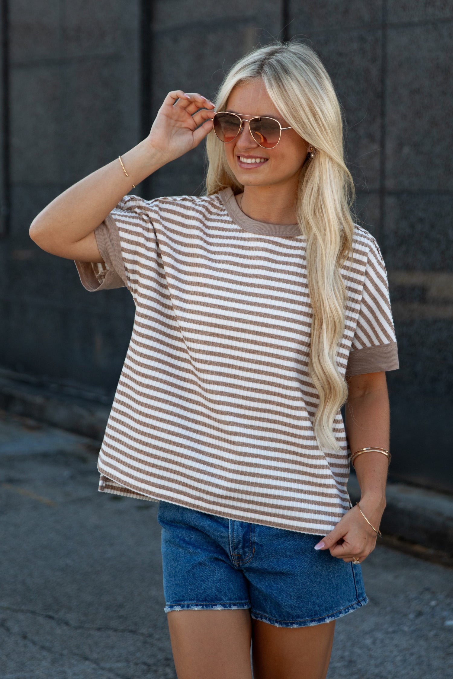 A woman wearing a brown and beige horizontally striped short-sleeve t-shirt with solid brown trim, paired with blue denim shorts, standing outdoors against a dark gray wall.