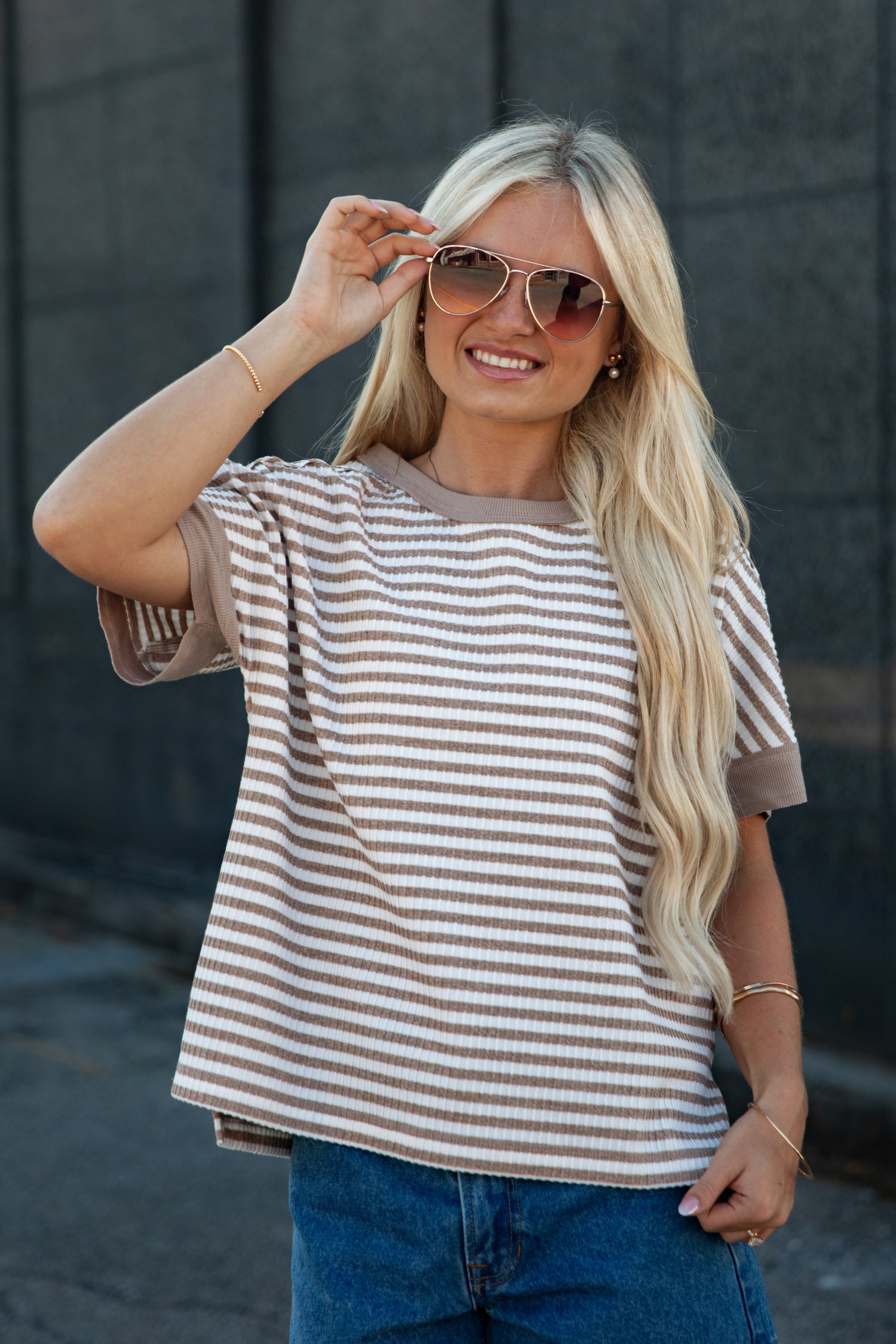 A woman with long blonde hair wearing a brown and beige horizontally striped short-sleeve t-shirt, blue jeans, and aviator sunglasses, posing against a dark gray brick wall.