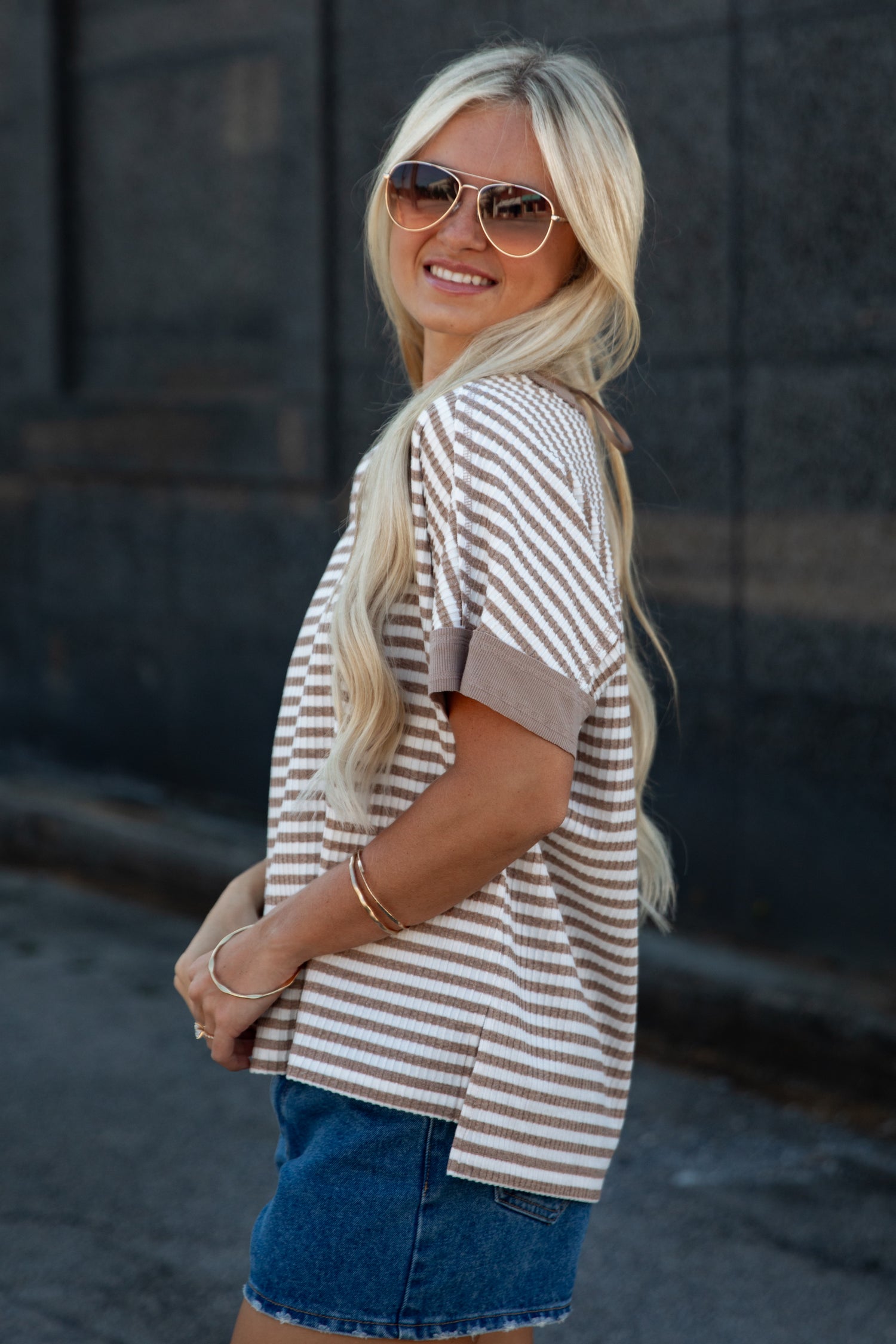 A woman with blonde hair and aviator sunglasses wearing a brown and beige striped short-sleeve top and denim shorts, posing outdoors against a dark stone wall.