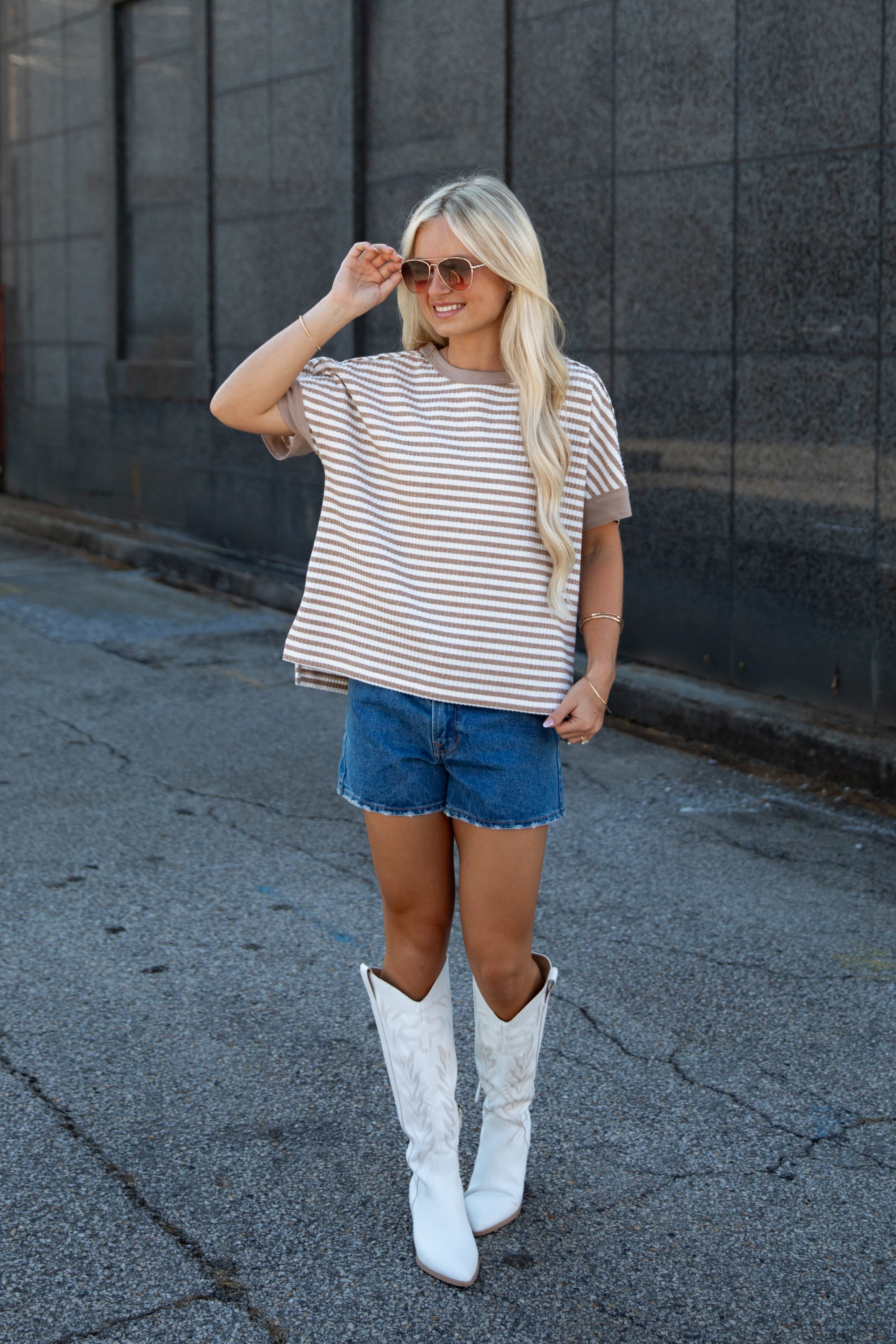A woman stands outdoors wearing a beige and white horizontally striped short-sleeve t-shirt, blue denim shorts, and tall white cowboy boots, posing in front of a dark gray stone wall.