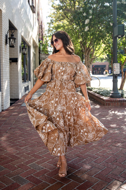 A woman wearing a brown off-the-shoulder maxi dress with a white floral pattern walks on a brick sidewalk in a sunny outdoor town setting.