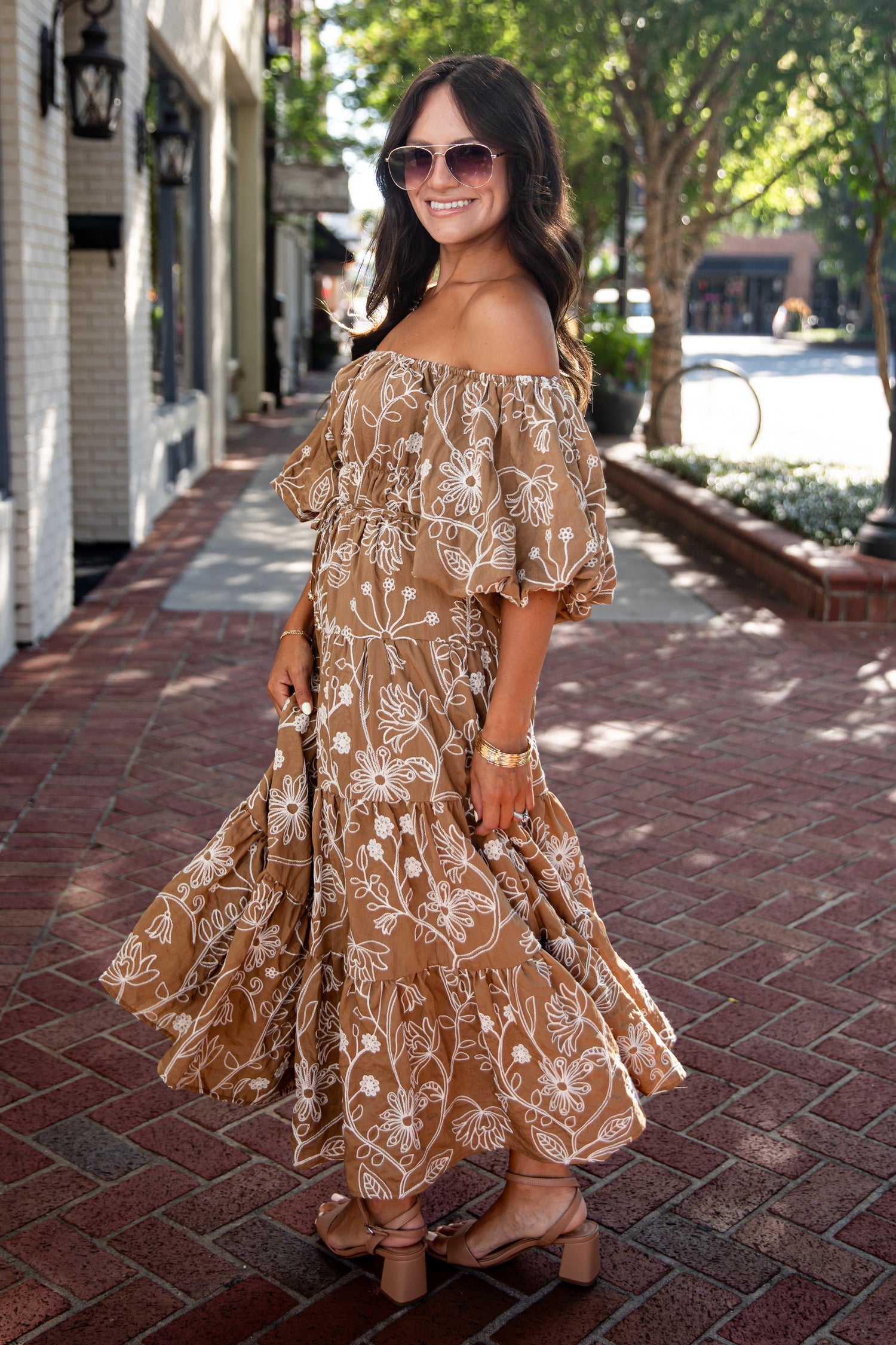 A woman wearing a brown off-the-shoulder maxi dress with a white floral pattern, standing on a red brick sidewalk in a town setting.