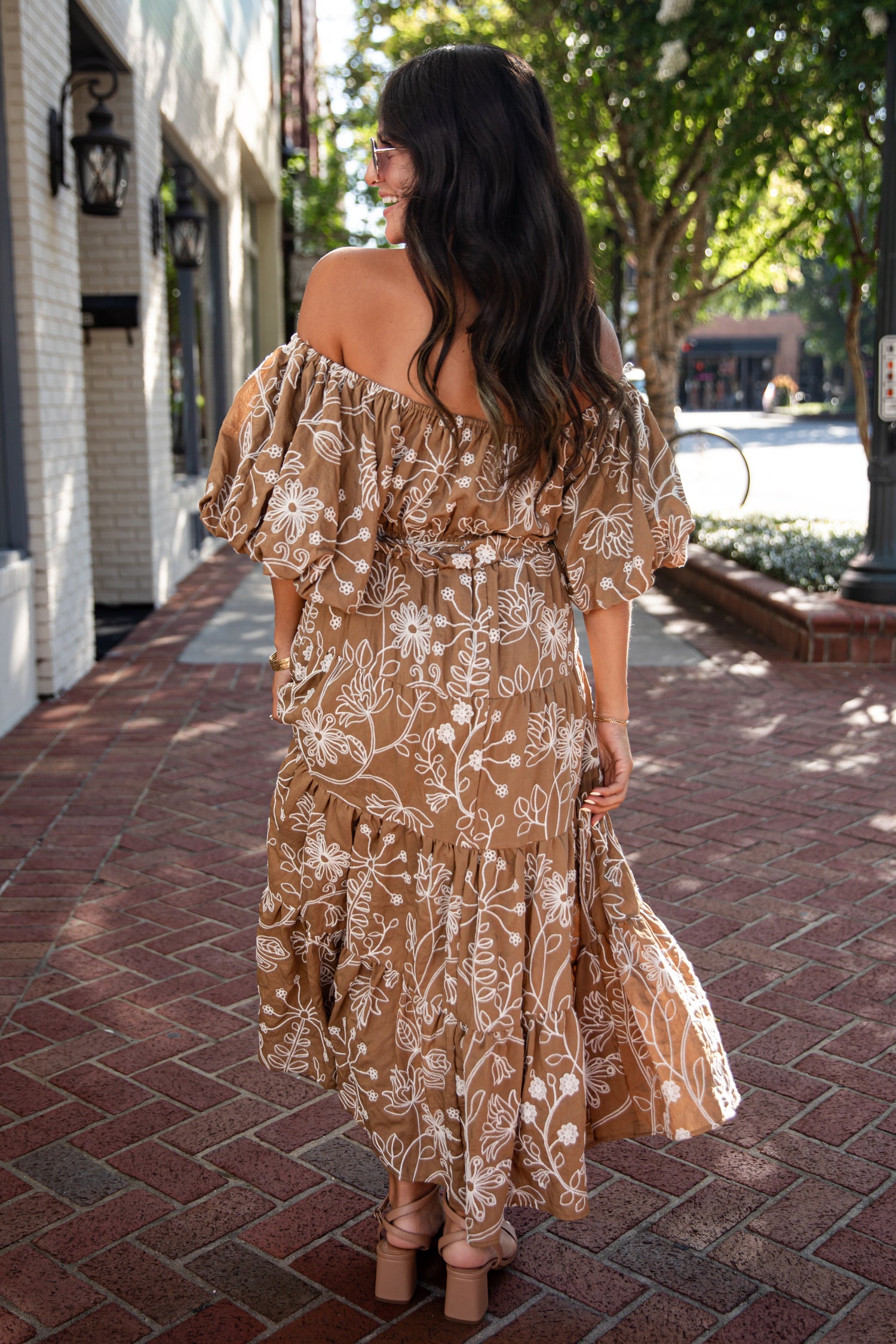 A woman viewed from behind wearing a brown off-the-shoulder maxi dress with a white floral pattern, walking on a brick sidewalk outdoors.