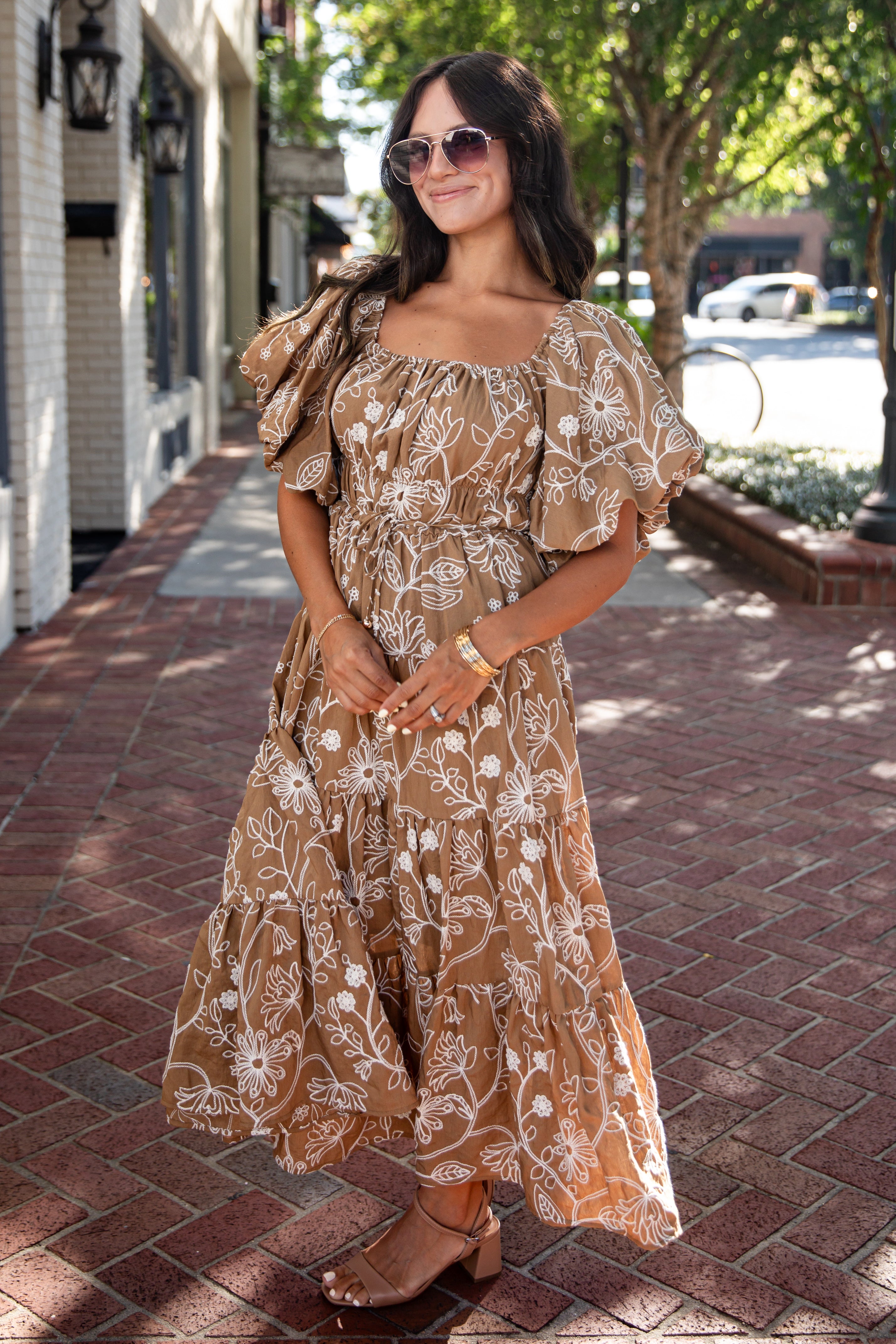 A woman wearing a brown and beige floral print maxi dress with puff sleeves, standing on a brick sidewalk in an outdoor street setting.