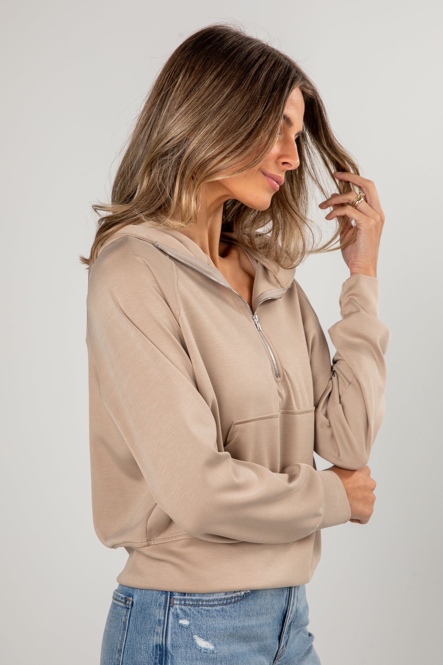 Woman wearing a beige zip-up hoodie and blue jeans against a plain background