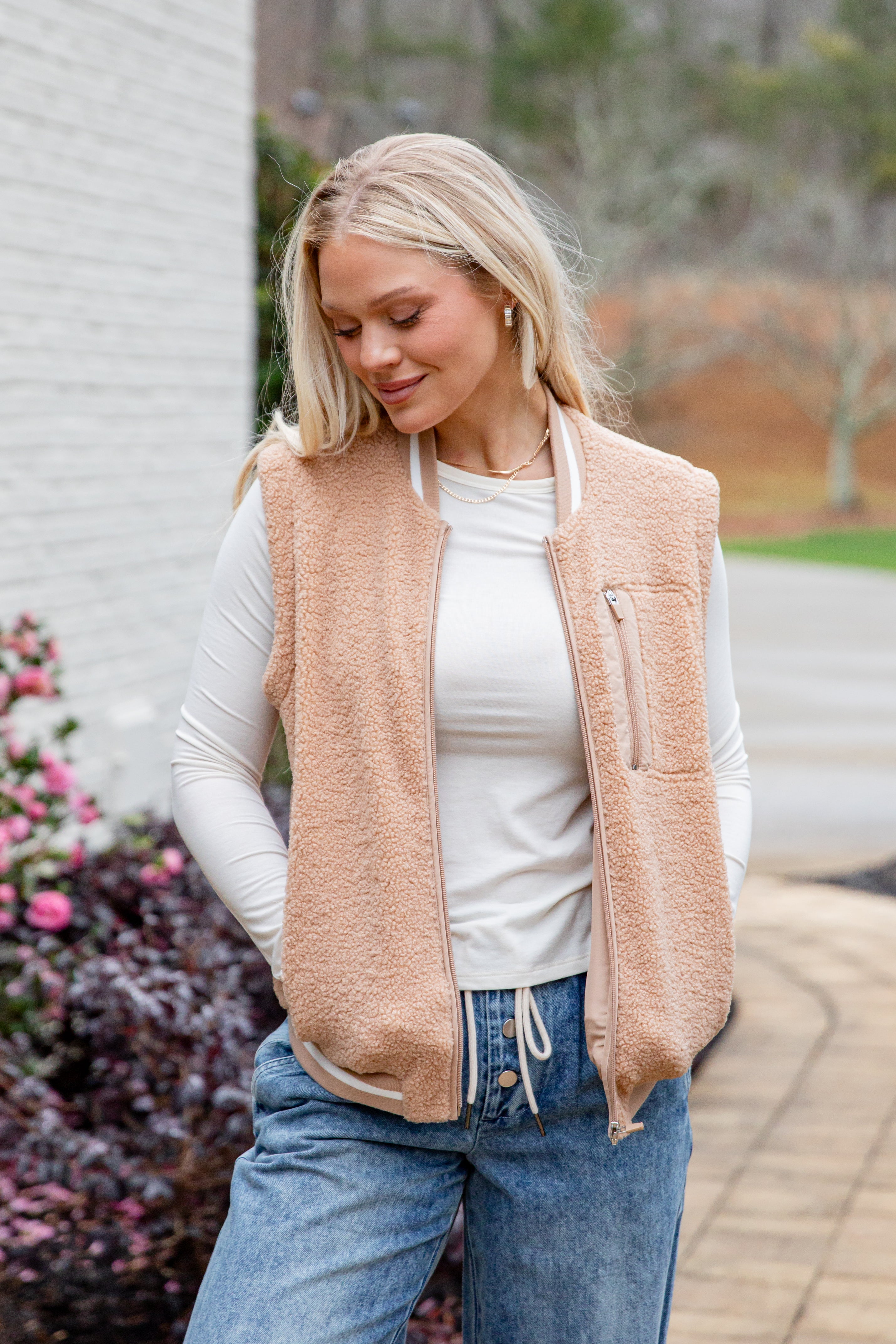 Woman wearing a beige vest over a white shirt and blue jeans, standing outdoors.