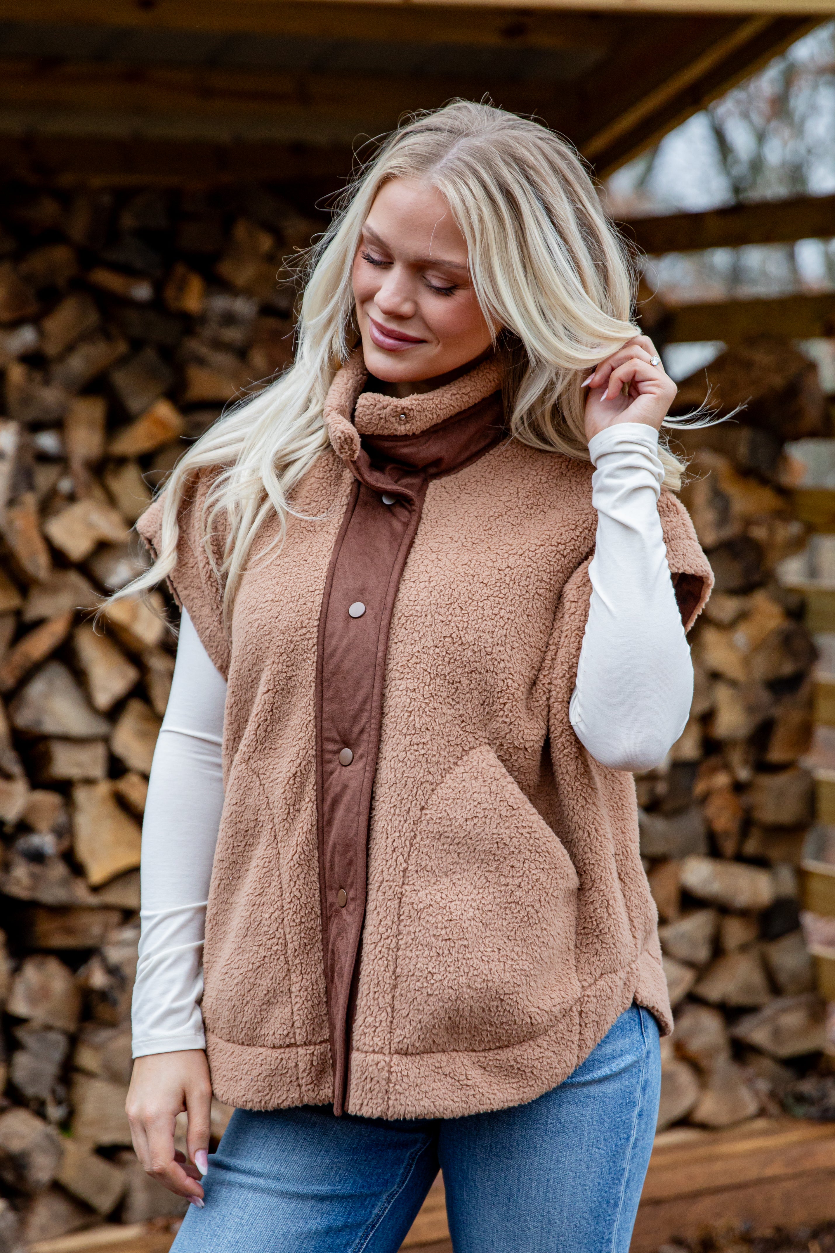 Woman wearing a brown vest over a brown shirt with a stack of firewood in the background