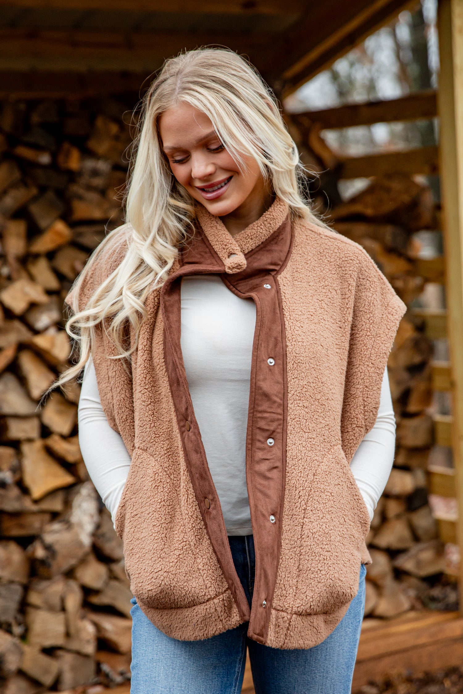 Woman wearing a brown vest over a white shirt in front of stacked firewood.