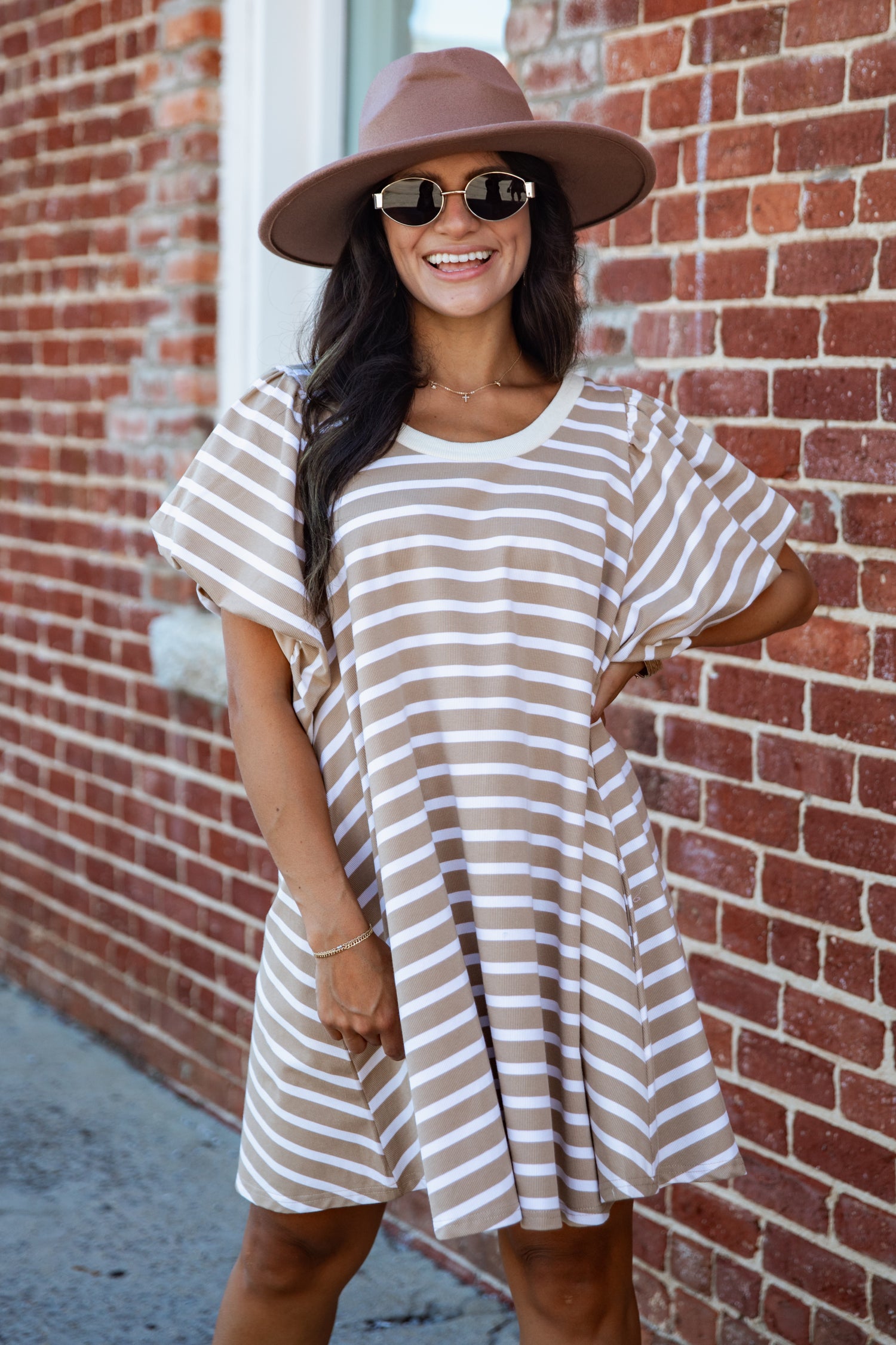 A woman wearing a brown and beige horizontally striped tunic dress, a brown wide-brim hat, and sunglasses, posing against a red brick wall.