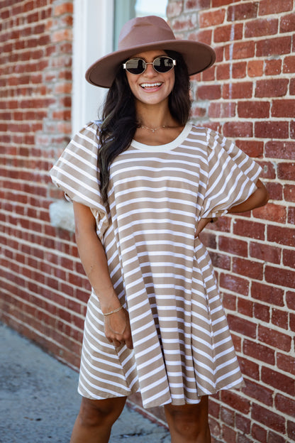 A woman wearing a brown and beige horizontally striped tunic dress, a brown wide-brim hat, and sunglasses, posing against a red brick wall.