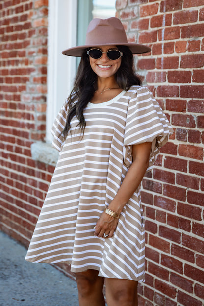 A woman wearing a beige and white horizontally striped short-sleeve tunic dress, a brown wide-brim hat, and round sunglasses, standing in front of a red brick wall.