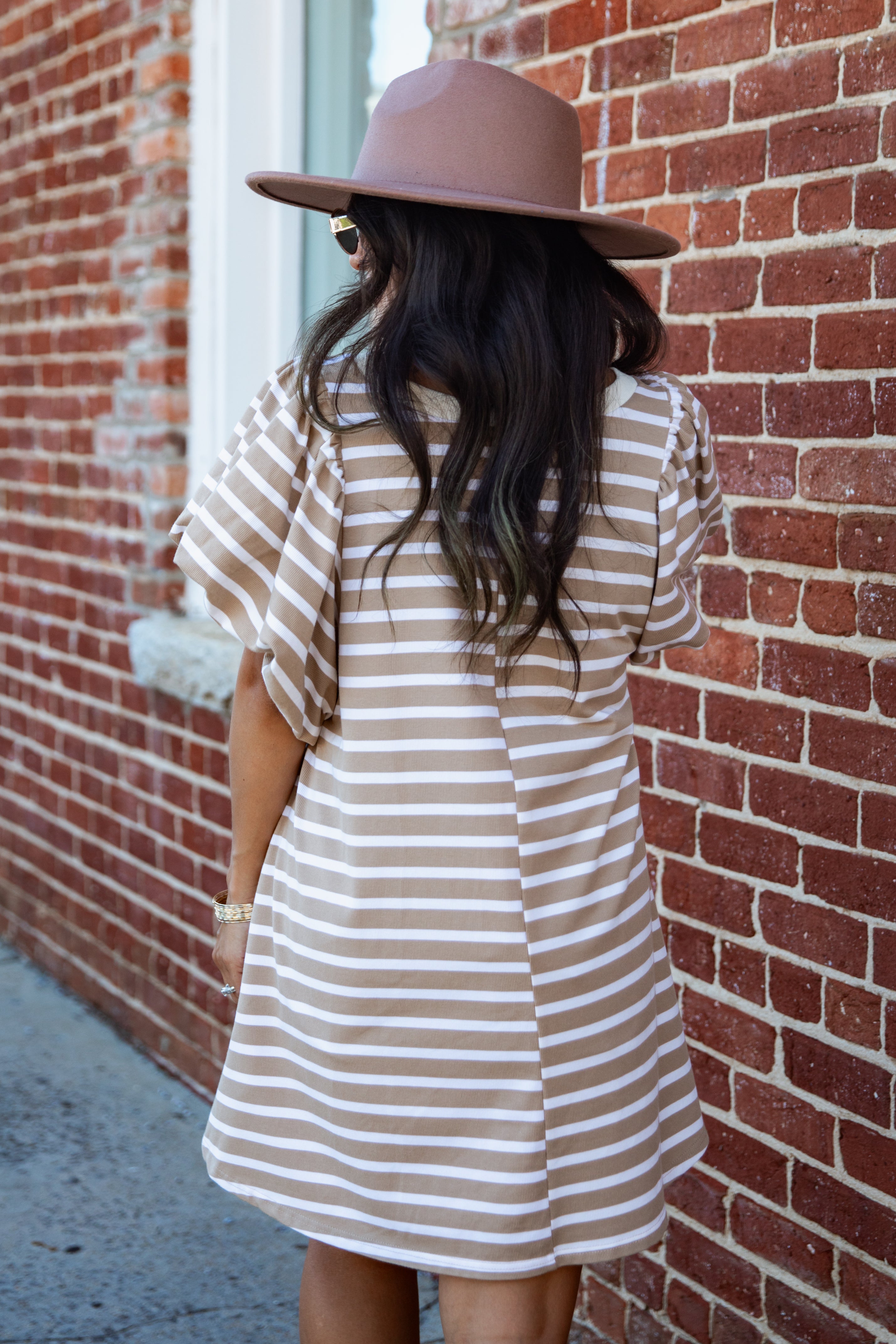 A woman viewed from behind wearing a brown wide-brim hat and a beige and white striped tunic dress, standing against a red brick wall.