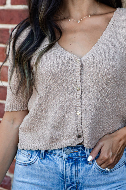 A woman wearing a short-sleeve knit cardigan top in a light taupe-beige color with gold-tone buttons, paired with light-wash blue jeans, standing in front of a red brick wall.