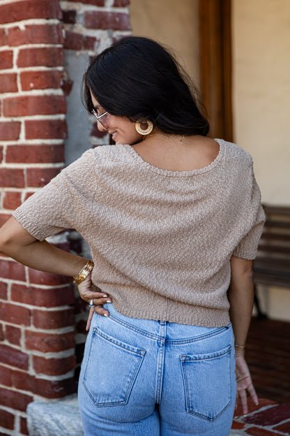 A woman wearing a beige short-sleeve knit top and light blue jeans, photographed from behind in an outdoor setting with a brick wall and wooden door in the background.