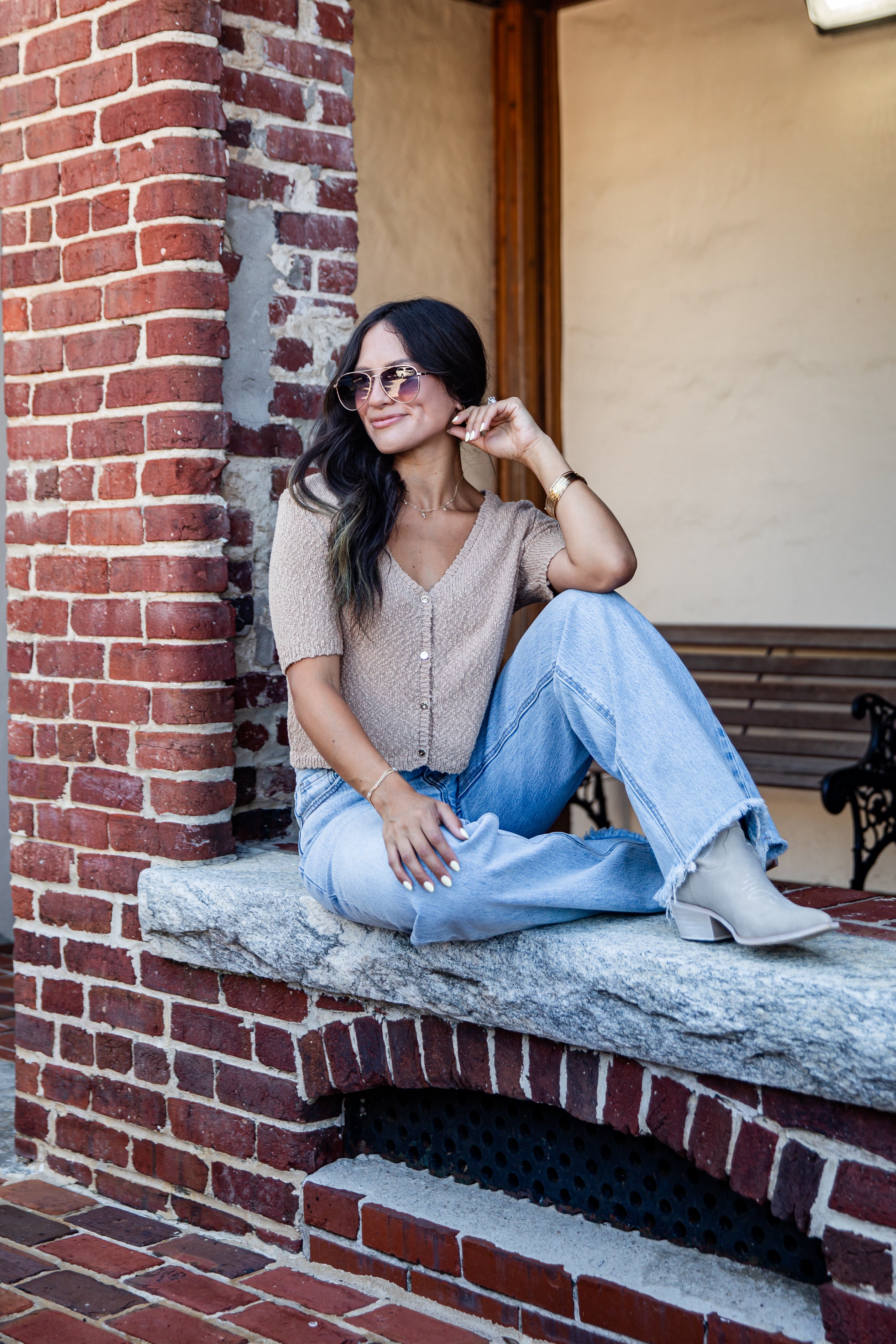 A woman sits on a stone ledge in front of a red brick column, wearing a beige knit short-sleeve top, light blue wide-leg jeans, and white heeled boots.