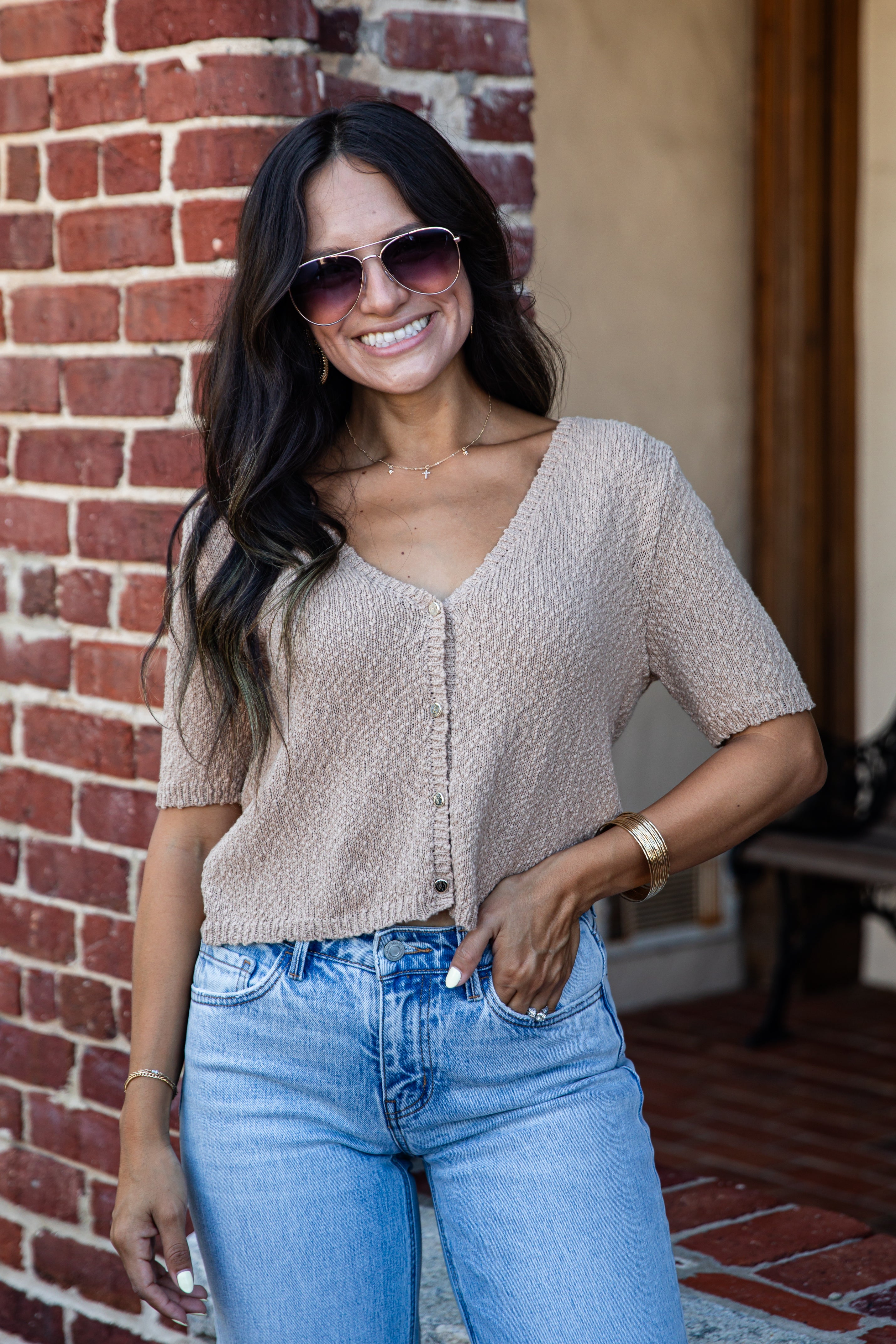 A woman wearing a short-sleeve beige textured knit top and light blue jeans, standing in front of a red brick wall.