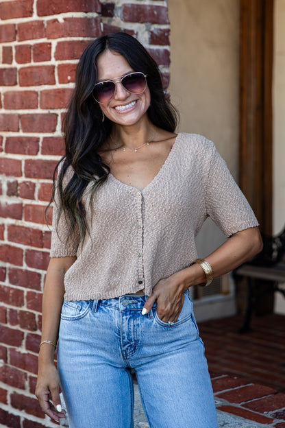 A woman wearing a short-sleeve beige textured knit top and light blue jeans, standing in front of a red brick wall.