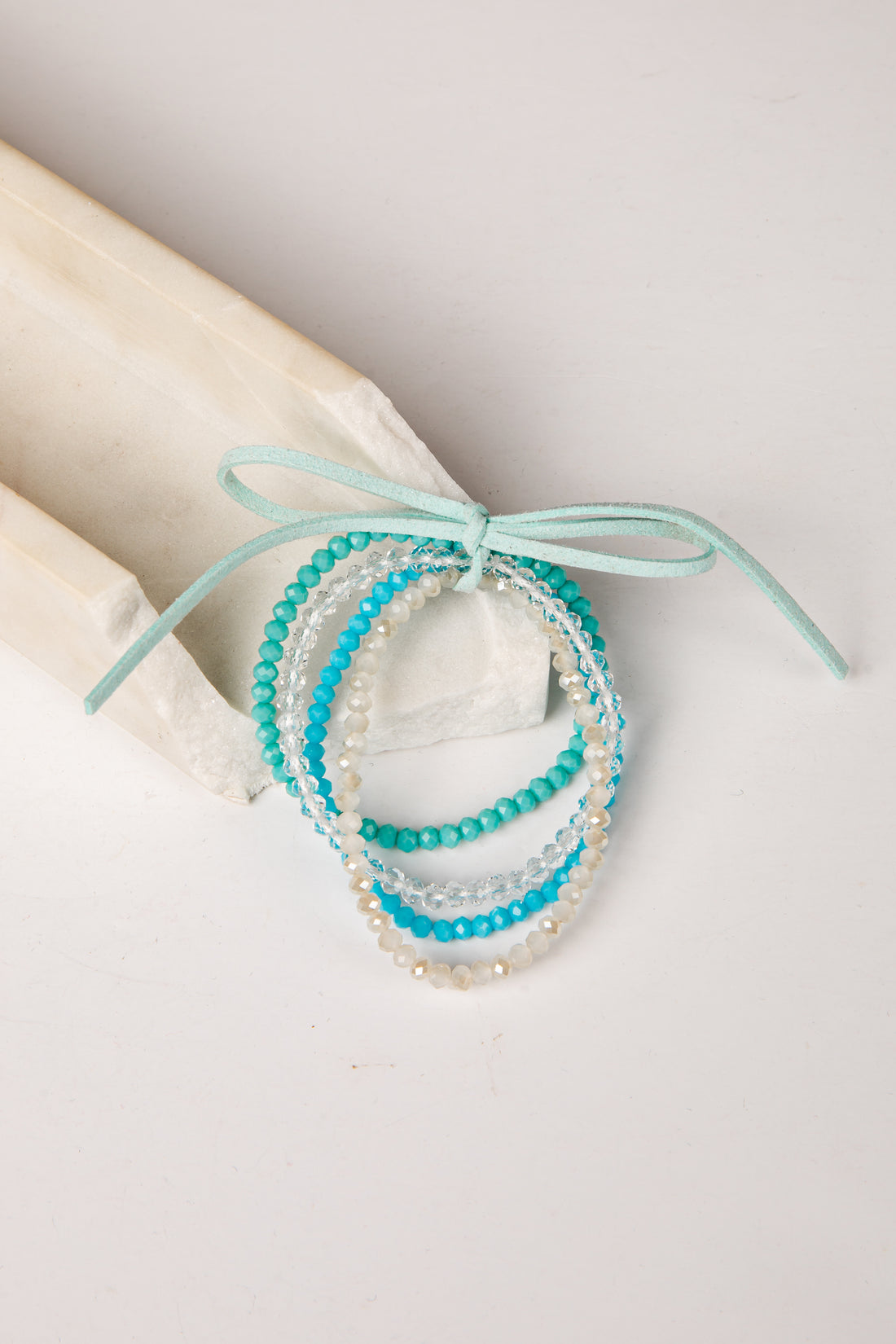 A set of three beaded stretch bracelets in turquoise, light blue, and white, tied together with a mint green ribbon on a light beige background.