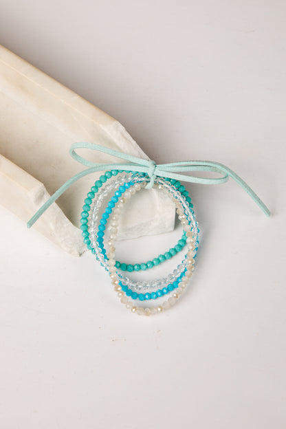 A set of three beaded stretch bracelets in turquoise, light blue, and white, tied together with a mint green ribbon on a light beige background.