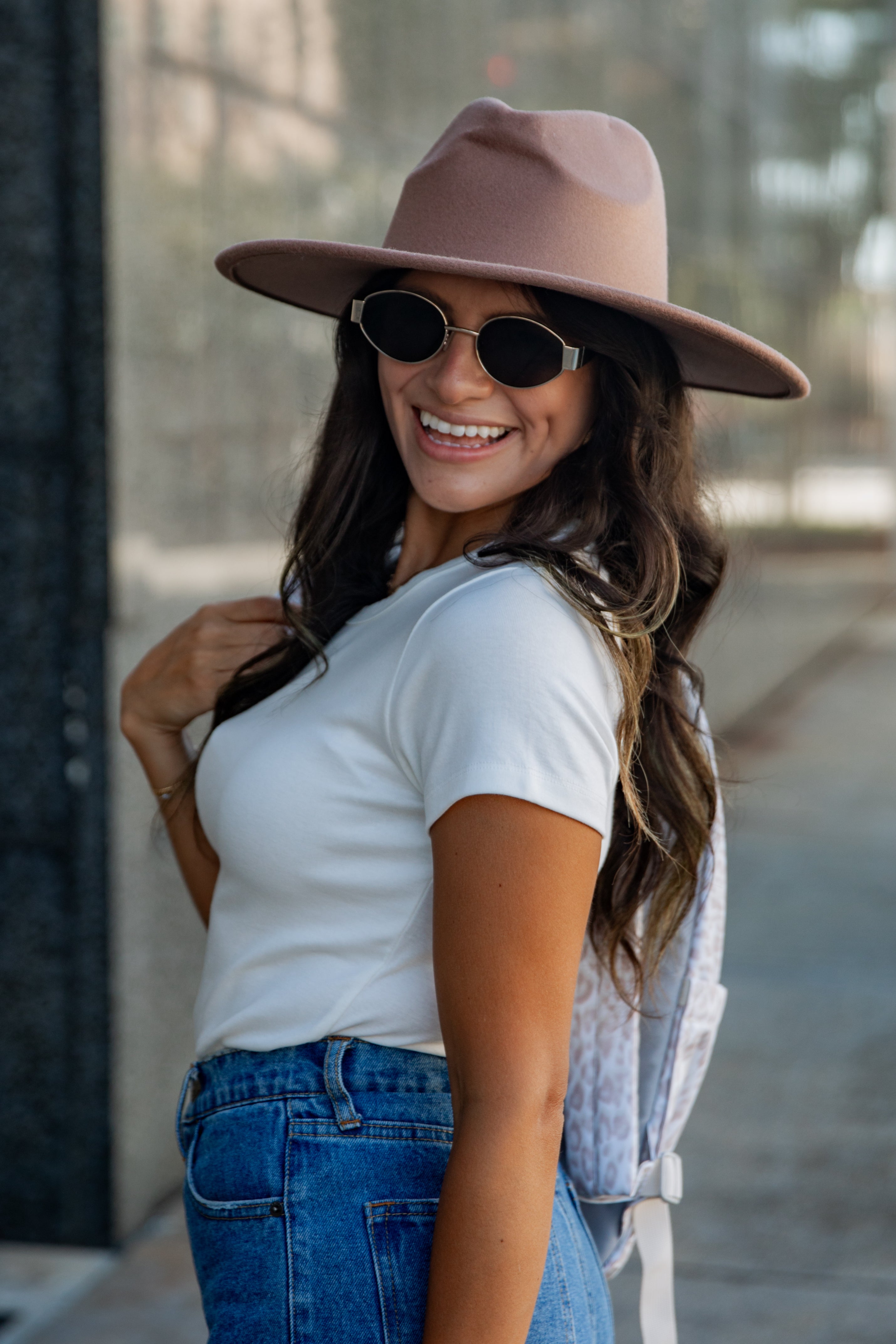 A woman wearing a dusty pink wide-brim fedora hat, round black sunglasses, a white t-shirt, and blue jeans, smiling outdoors on a city sidewalk.