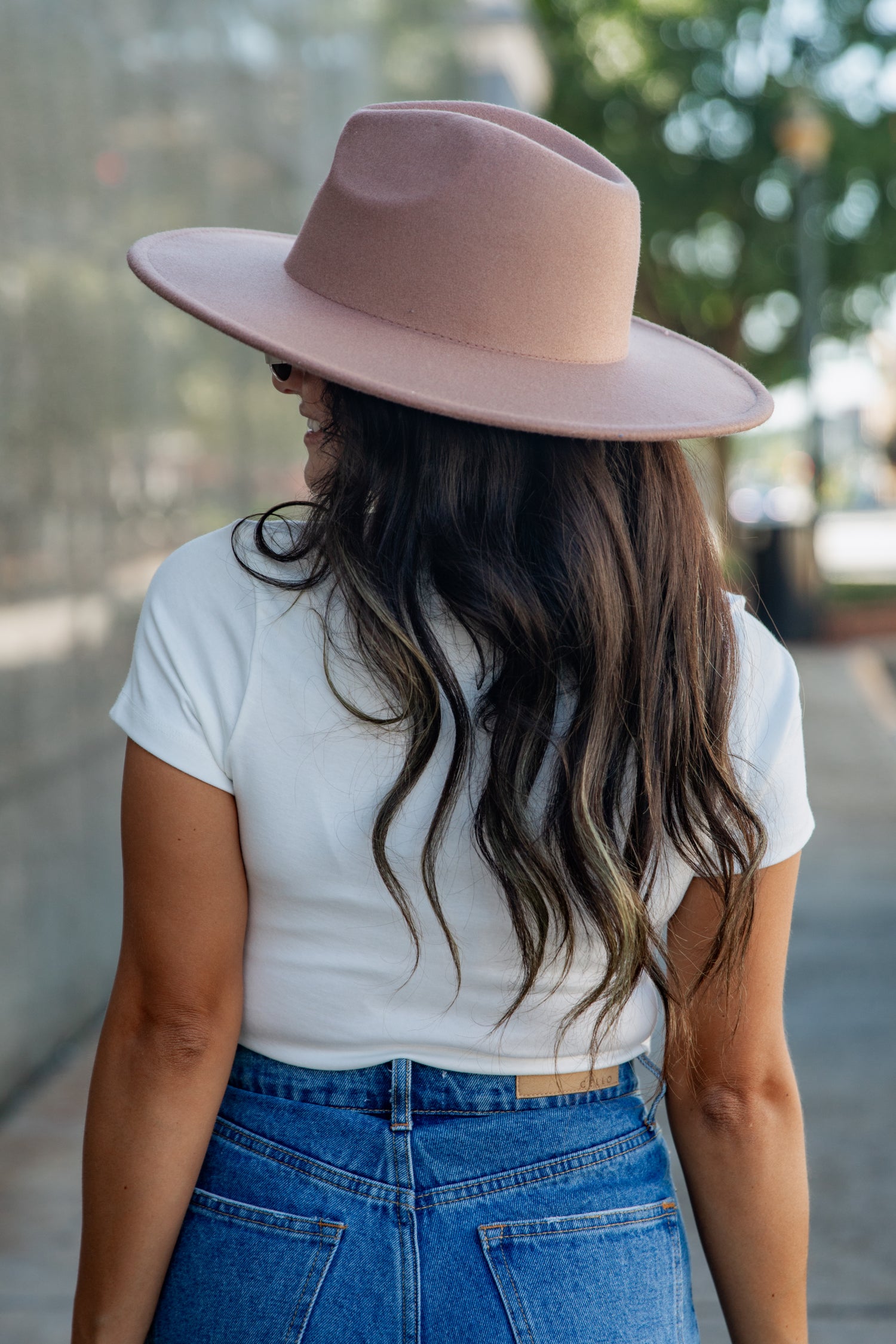 A woman viewed from behind wearing a dusty pink wide-brim fedora hat, a white t-shirt, and blue jeans, set against a softly blurred outdoor background.