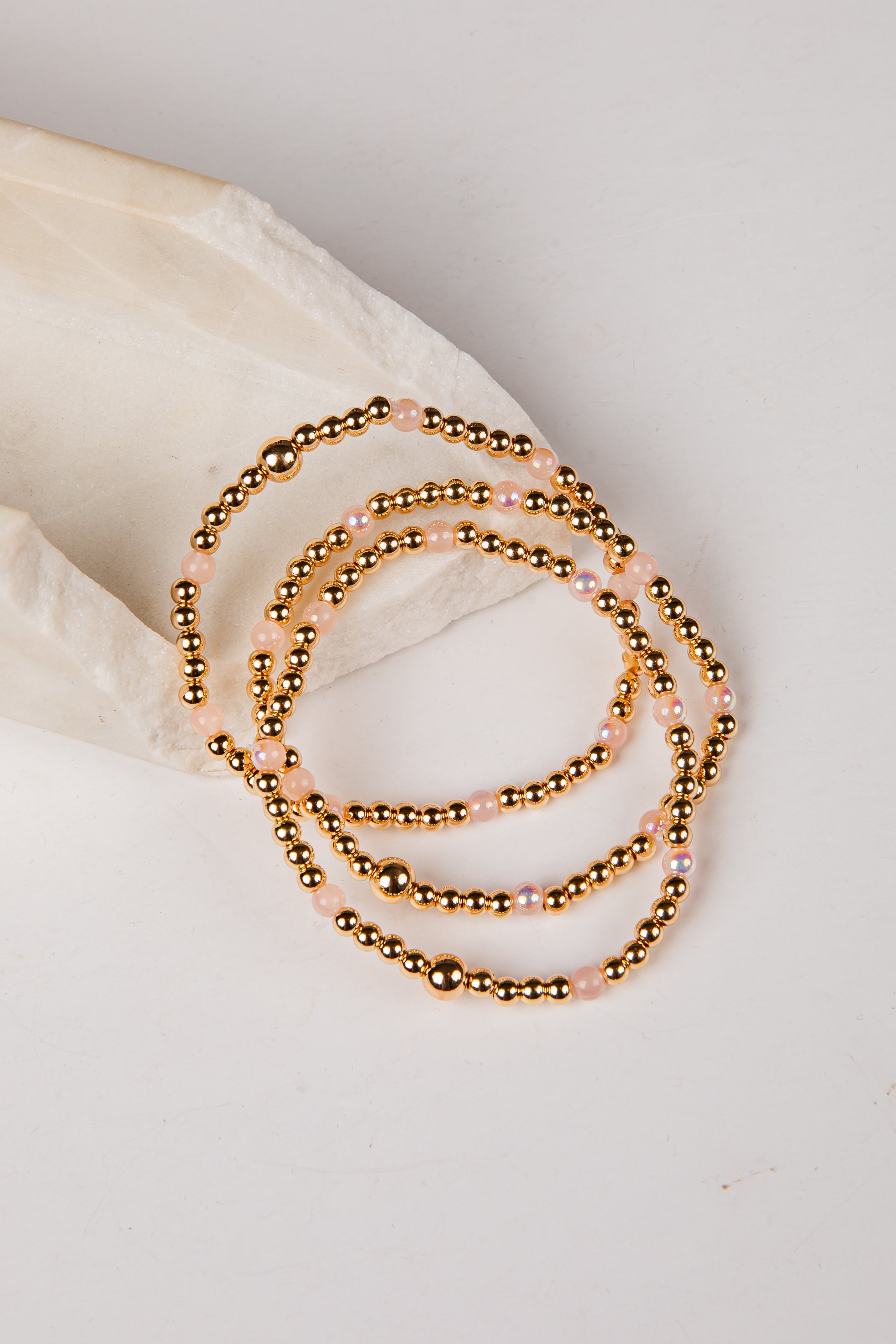 A stack of three beaded stretch bracelets with metallic gold beads and small iridescent pink beads, arranged on a light cream fabric prop against a white background.