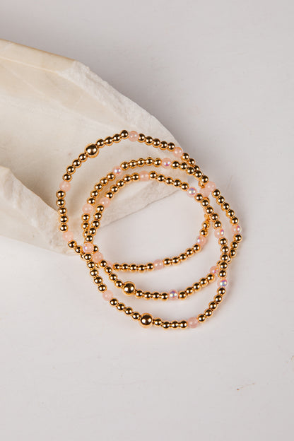 A stack of three beaded stretch bracelets with metallic gold beads and small iridescent pink beads, arranged on a light cream fabric prop against a white background.