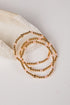 A stack of three beaded stretch bracelets with metallic gold beads and small iridescent pink beads, arranged on a light cream fabric prop against a white background.