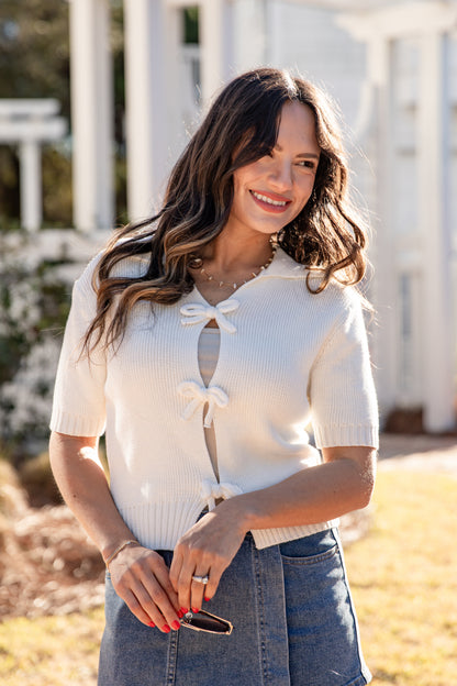 Woman wearing a white short-sleeve top with a bow detail, standing outdoors.