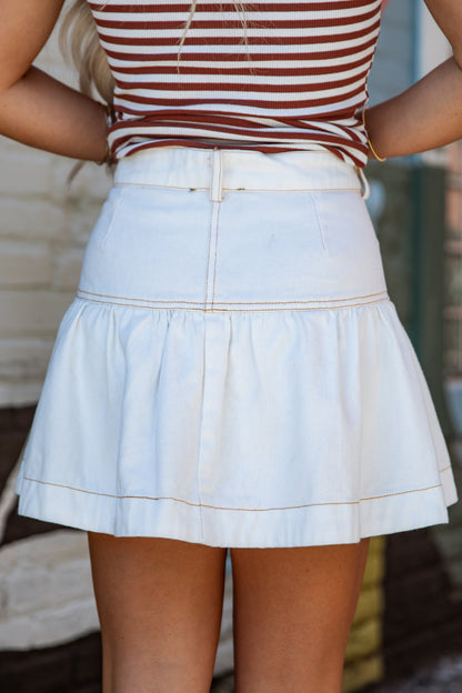 A person wearing a white tiered mini skirt with thin gold contrast stitching and a matching white belt, paired with a brown and white striped top.