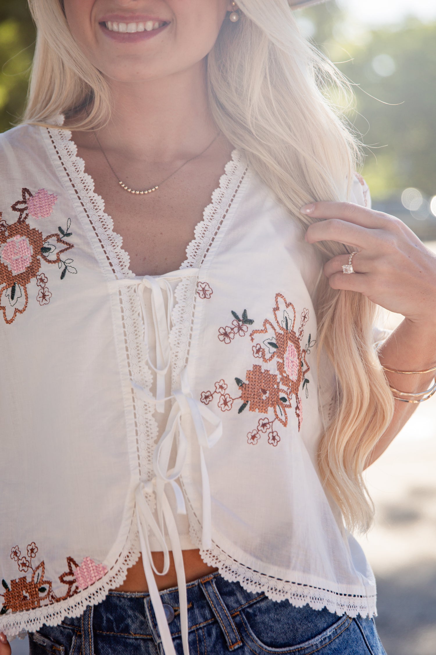 A close-up of a woman wearing a white lace-up crop top with pink and brown floral embroidery, paired with blue jeans in an outdoor setting.