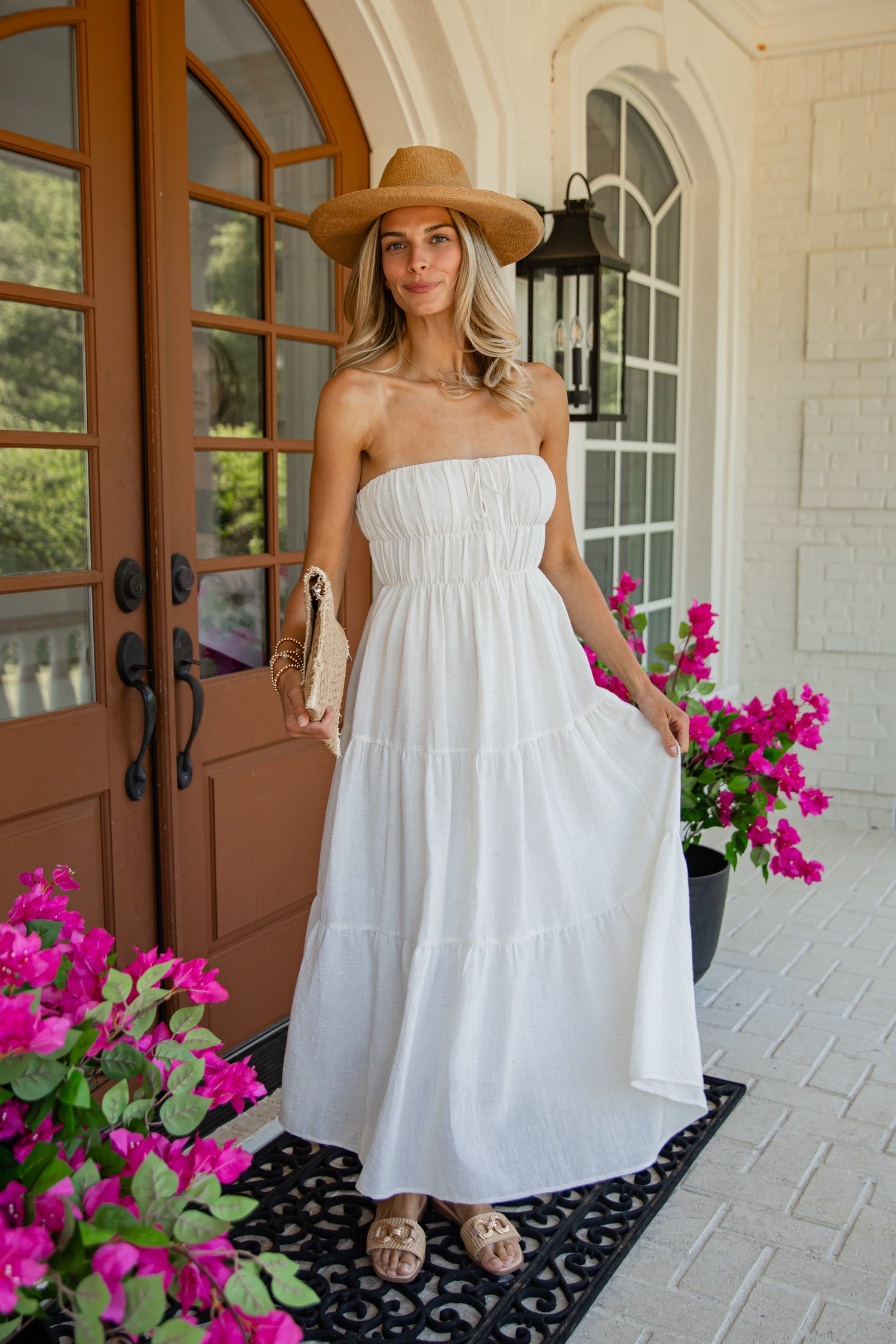 A woman stands on a front porch wearing a strapless white tiered maxi dress, a tan wide-brim hat, and tan sandals. Bright pink flowers and a brown door are visible in the background.