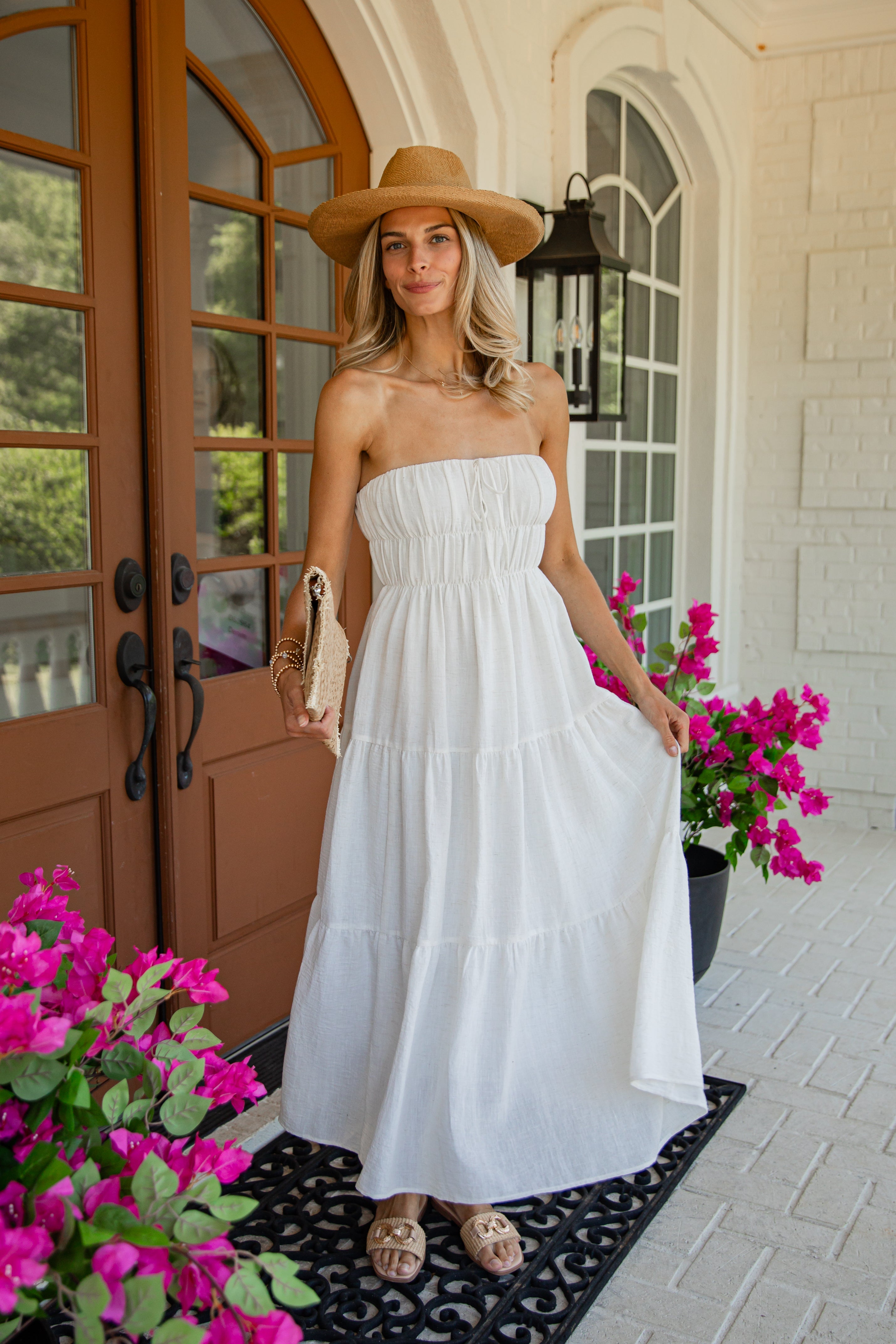 A woman stands on a front porch wearing a strapless white tiered maxi dress, a tan wide-brim hat, and tan sandals. Bright pink flowers and a brown door are visible in the background.