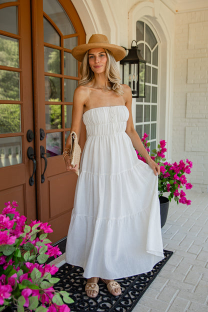 A woman stands on a front porch wearing a strapless white tiered maxi dress, a tan wide-brim hat, and tan sandals. Bright pink flowers and a brown door are visible in the background.