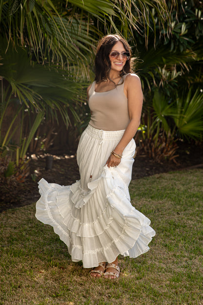 Woman in a beige top and white ruffled skirt standing outdoors with greenery in the background