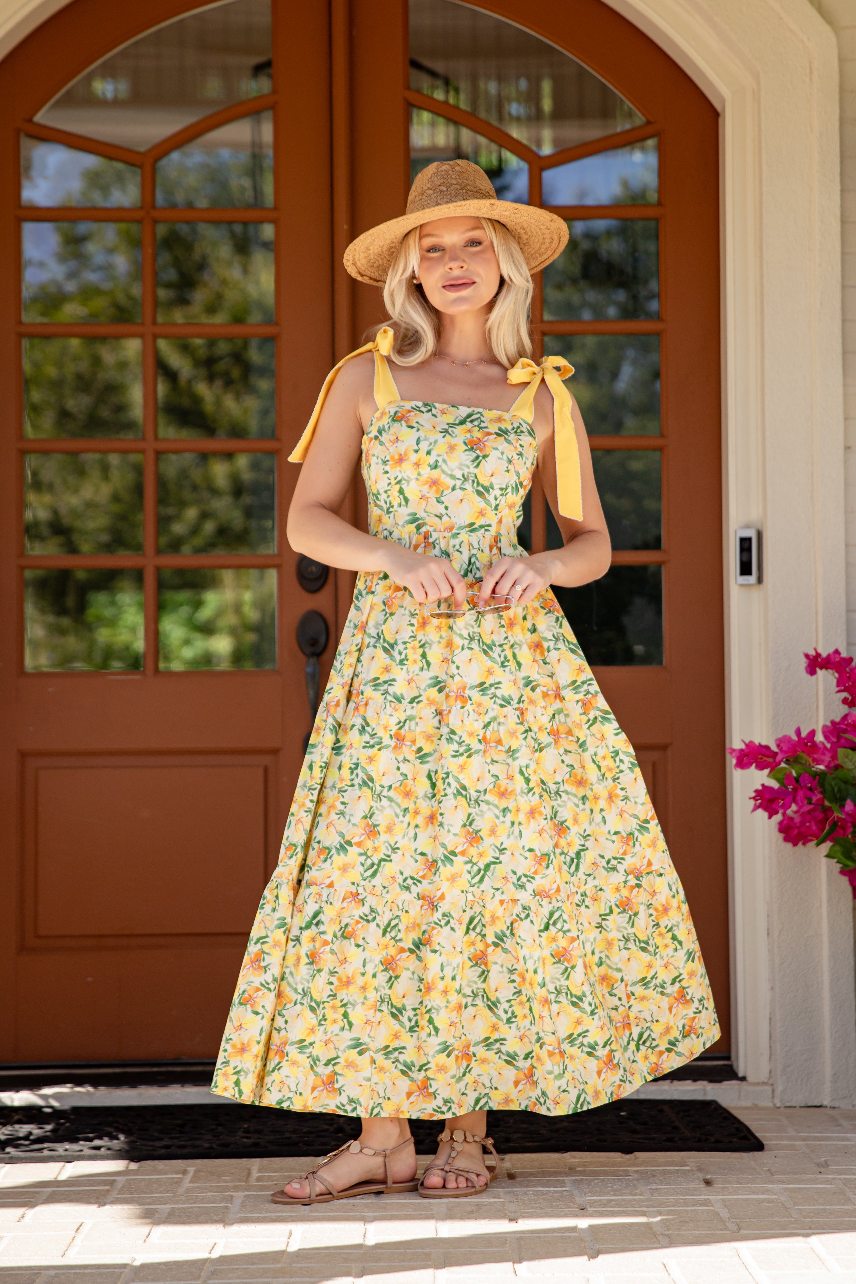 Woman in a floral dress and sun hat standing in front of a brown door with glass panels.
