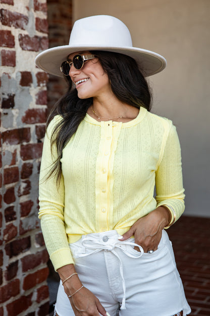 A woman wearing a pale yellow ribbed knit cardigan, white shorts, a white wide-brim hat, and sunglasses, standing in front of a brick wall.