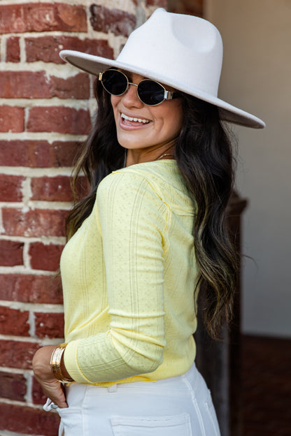A woman wearing a white wide-brim fedora hat, round sunglasses, a pale yellow ribbed knit long-sleeve top, and white pants, posing in front of a red brick wall.