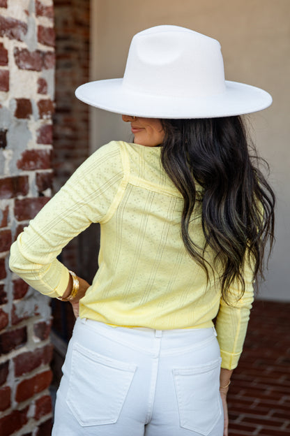 A woman shown from the back wearing a pale yellow textured knit top, white pants, and a large white wide-brim hat, standing outdoors next to a brick wall.