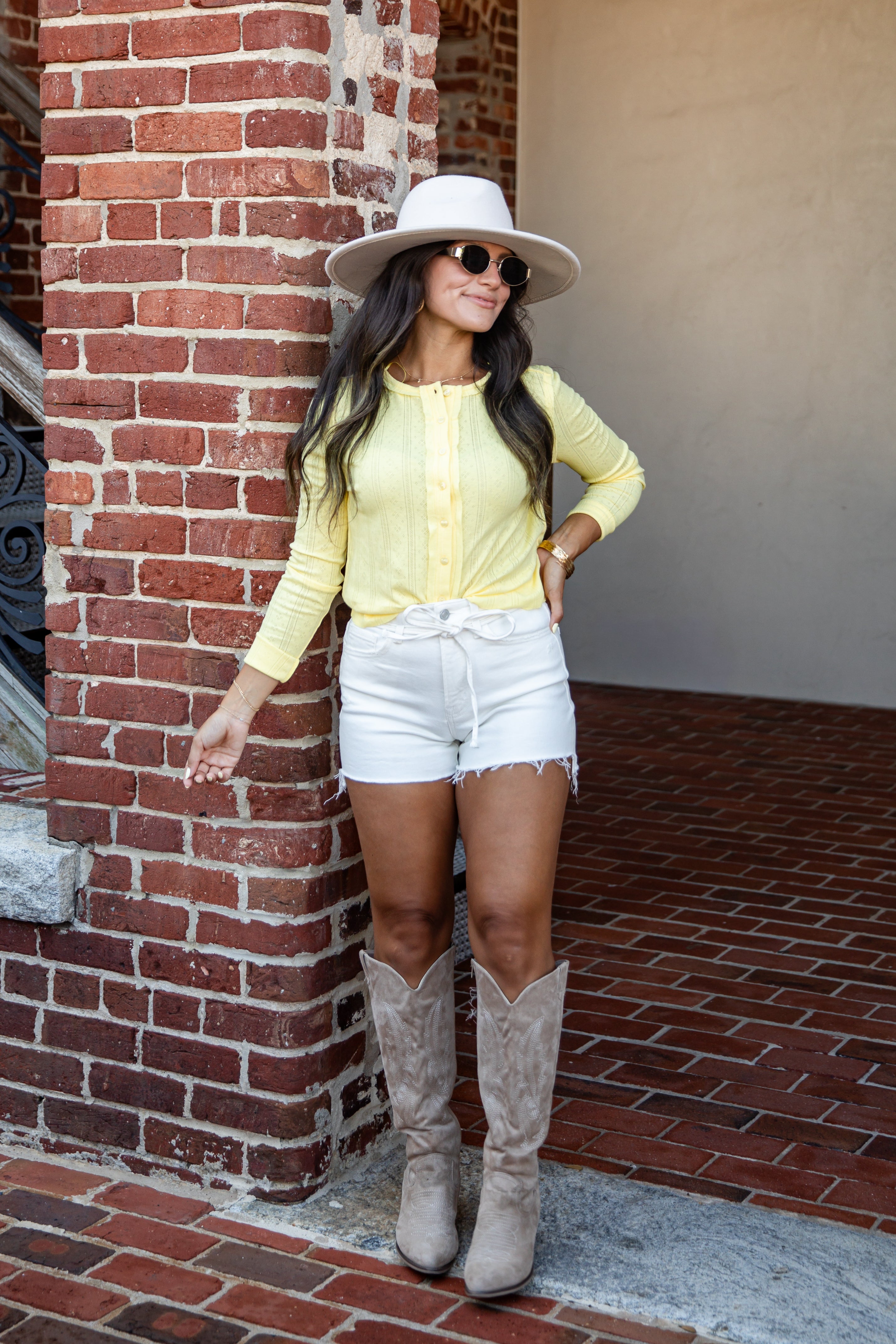 A woman posing in a yellow long-sleeve button-up shirt, white denim shorts, a white wide-brim hat, and beige cowboy boots, standing next to a red brick column.