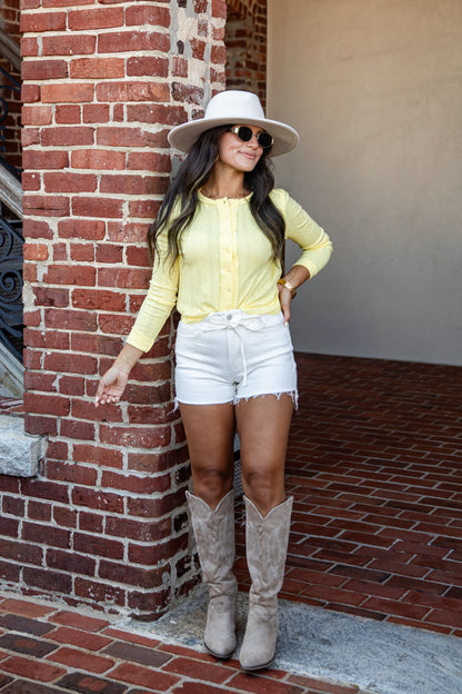 A woman posing in a yellow long-sleeve button-up shirt, white denim shorts, a white wide-brim hat, and beige cowboy boots, standing next to a red brick column.