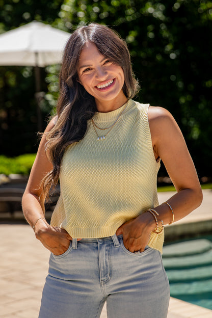 Woman wearing a yellow sleeveless sweater and blue jeans standing outdoors.
