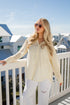 Woman in a yellow blouse and white pants standing on a balcony with a coastal background